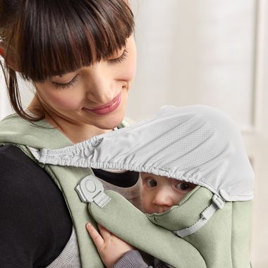 A woman smiles while carrying an infant in a green baby carrier. The baby, partially covered by a white canopy, gazes curiously.