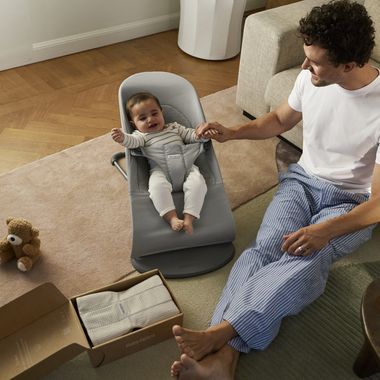 A baby in a gray seat smiles while holding hands with an adult sitting on the floor, with a teddy bear and an open box nearby.