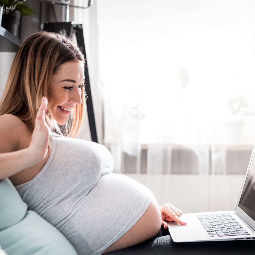 Donna incinta seduta sul divano, sorride e saluta guardando uno schermo di laptop in un ambiente domestico luminoso.