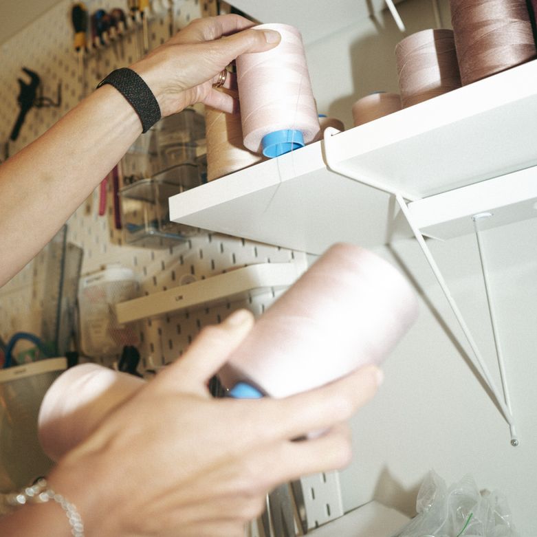 Hands placing pink thread spools on a white shelf with a pegboard wall in the background holding various tools.