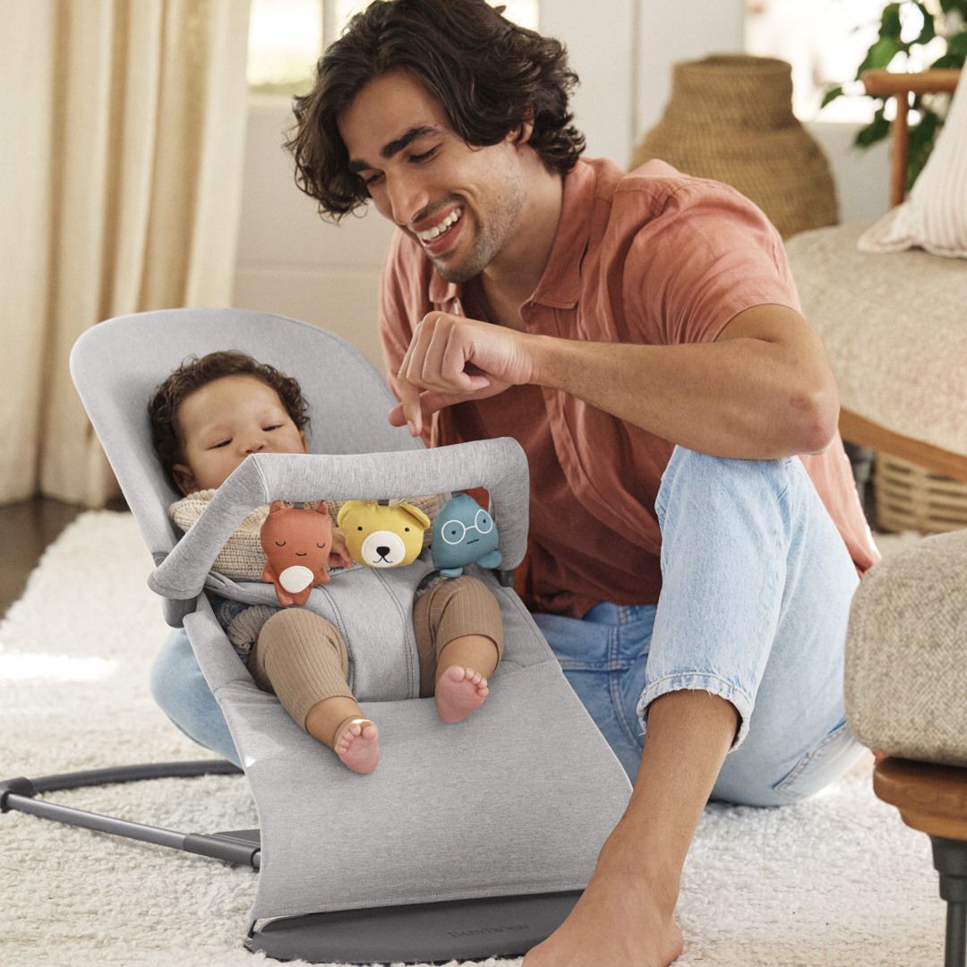 A smiling man interacts with a baby in a gray baby bouncer, surrounded by plush toys. The room is cozy with soft lighting.