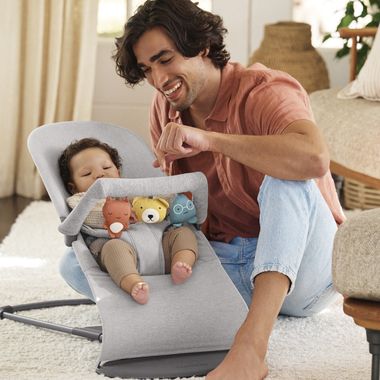 A smiling man interacts with a baby in a gray baby bouncer, surrounded by plush toys. The room is cozy with soft lighting.