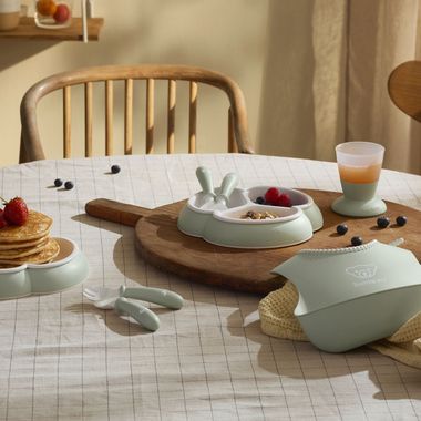 Children's table setting with pancakes, fruit, utensils, and a bib in light green. Juice in a cup, on a checkered tablecloth.