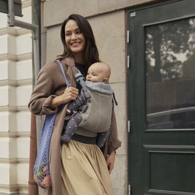 Woman walking with a baby in a carrier, wearing a long coat and skirt, holding a mesh bag with groceries, smiling on a city sidewalk.