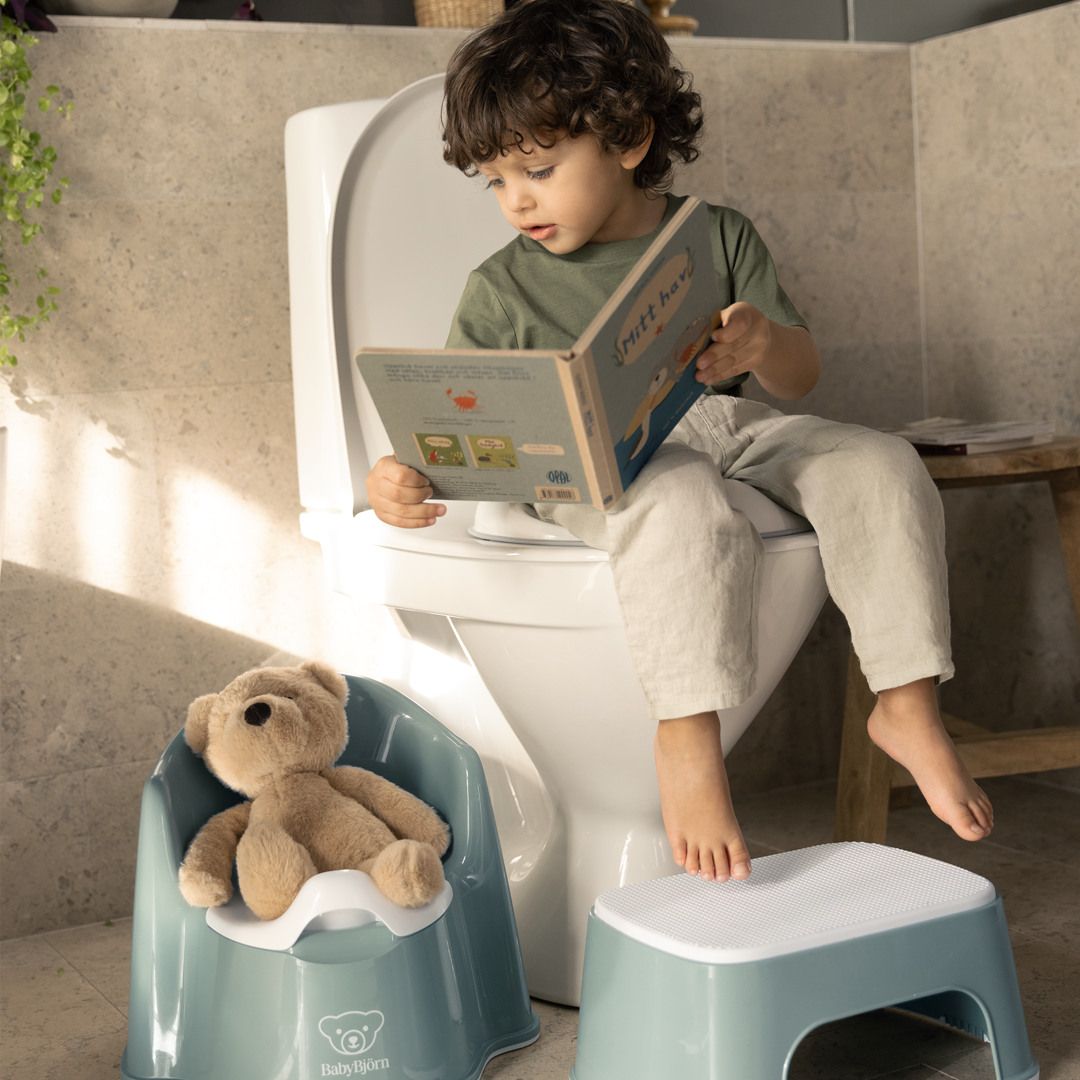 Child sitting on a toilet reading a book, with a teddy bear on a training potty and a step stool nearby, in a sunlit bathroom.