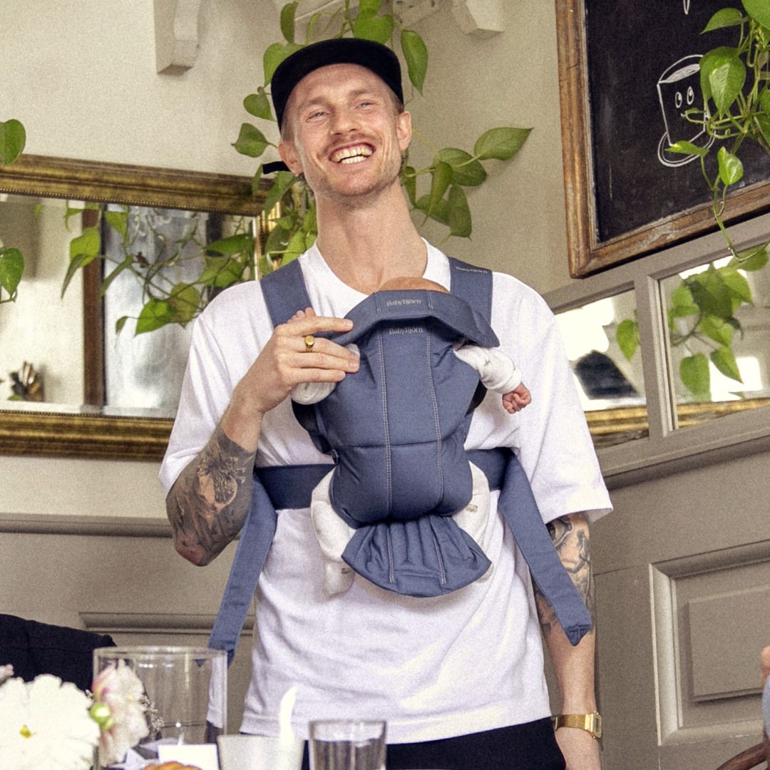 Smiling man with tattoos and a cap carries a baby in a front baby carrier, surrounded by plants and framed pictures in a cozy indoor setting.
