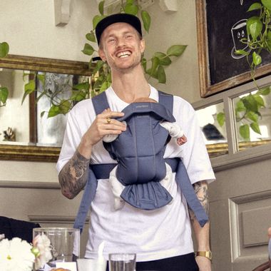Smiling man with tattoos and a cap carries a baby in a front baby carrier, surrounded by plants and framed pictures in a cozy indoor setting.