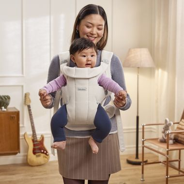 A woman smiling as she holds a baby in a carrier inside a cozy living room with a guitar and a lamp in the background.