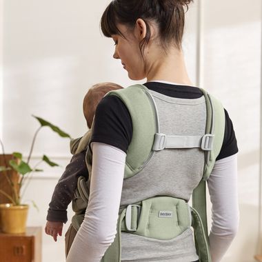 Woman wearing a baby in a green carrier, standing indoors near a potted plant.