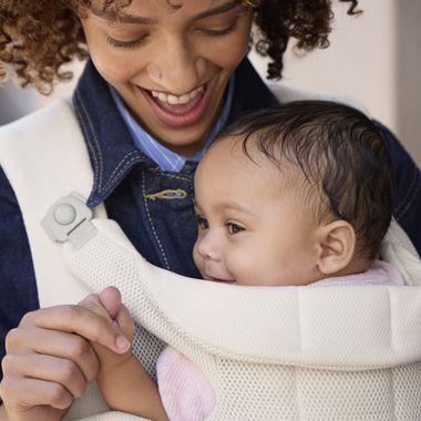 A smiling woman in a denim jacket holds a baby in a light-colored baby carrier. The baby looks content, holding the woman's finger.