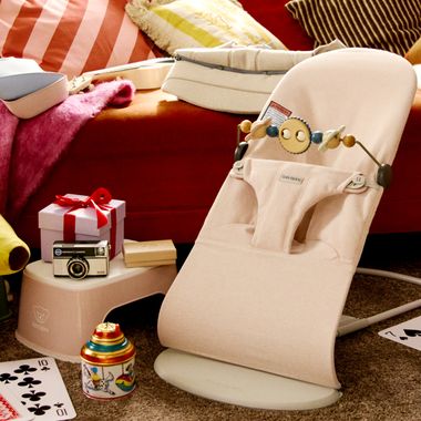 A beige baby bouncer with a toy bar sits on a carpet. Nearby are a gift box, vintage camera, colorful toy, and playing cards, all against a striped sofa.