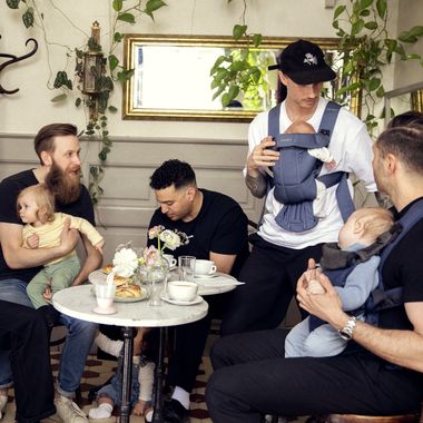 Group of men with babies, sitting and interacting at a café table. Plants and mirrors in the background. Casual, friendly atmosphere.