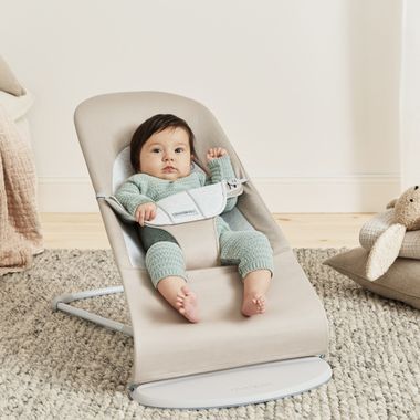 A baby in a light green outfit sits in a beige baby bouncer on a carpeted floor, with soft toys nearby.
