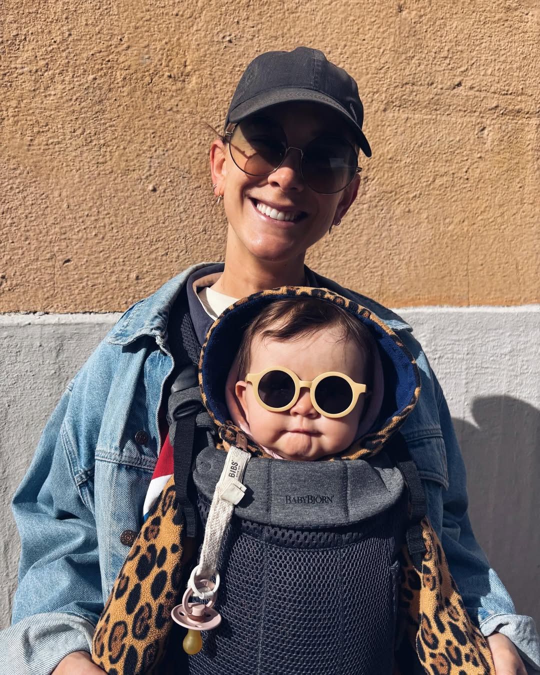 Smiling person in a denim jacket and cap carrying a baby in a leopard-print outfit and sunglasses in a front carrier.