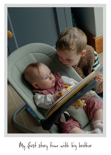 A young child reads a book to a baby sitting in a rocker, with soft toys around. The baby looks engaged and attentive.
