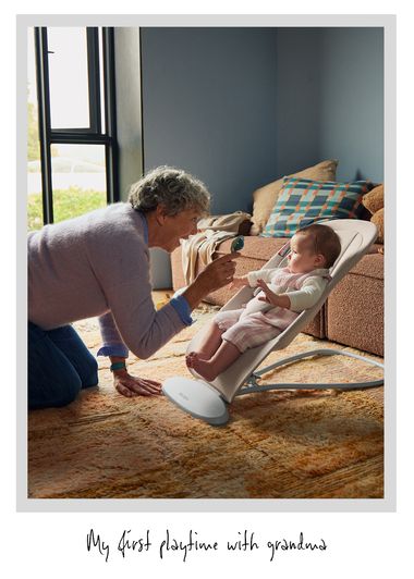 An elderly woman plays with a baby seated in a bouncer in a cozy living room with soft lighting.