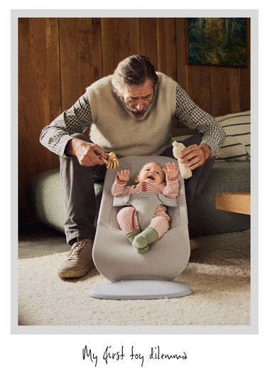 An elderly man presents a baby in a bouncer with a wooden toy in one hand and a stuffed toy in the other, in a cozy wooden room.