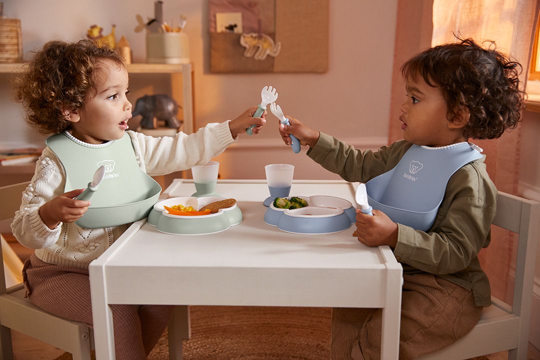 Two toddlers sit at a small table, wearing bibs, playfully holding utensils. Plates with food and cups are in front of them.