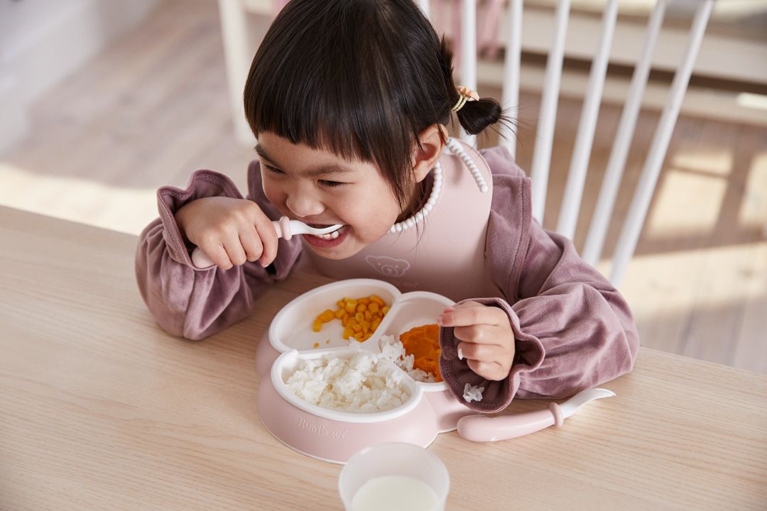 Young child eating from a divided plate with rice, corn, and a vegetable. Wearing a bib and using a spoon from BabyBjörn