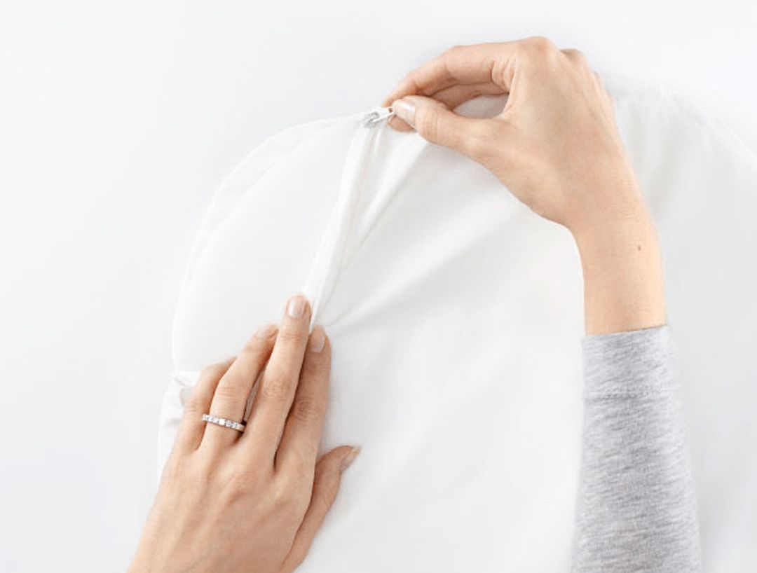 Hands unzipping a white fabric cover, revealing part of the material inside. One hand wears a ring. Gray sleeve visible on one arm.