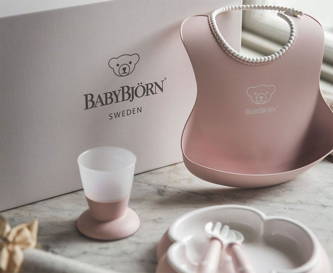 Pink BabyBjörn feeding set with bib, cup, and divided plate on a table. Background shows a branded box with a bear logo.