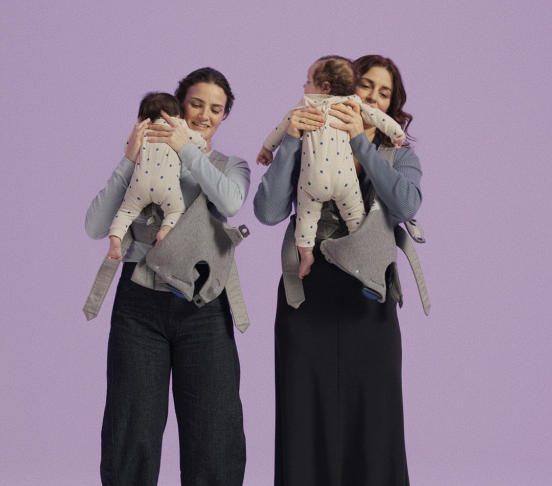 Two women hold babies with star-patterned onesies, using grey baby carriers against a lavender background.