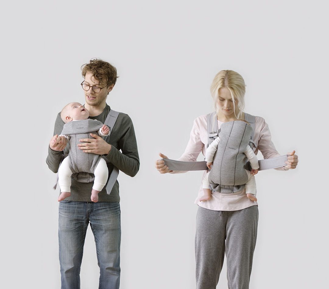Two adults, each wearing a baby carrier with an infant, demonstrate how to use the carriers. The background is light gray.