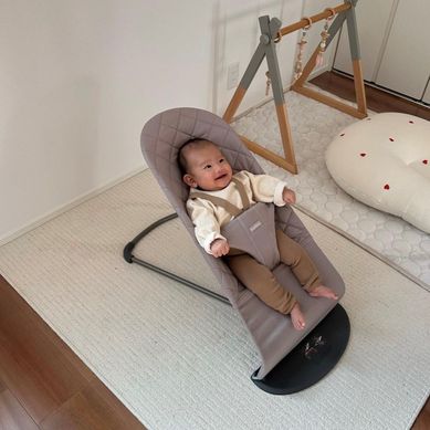 A baby sits smiling in a gray bouncer on a cream rug, with a play gym and cushion nearby in a cozy room.