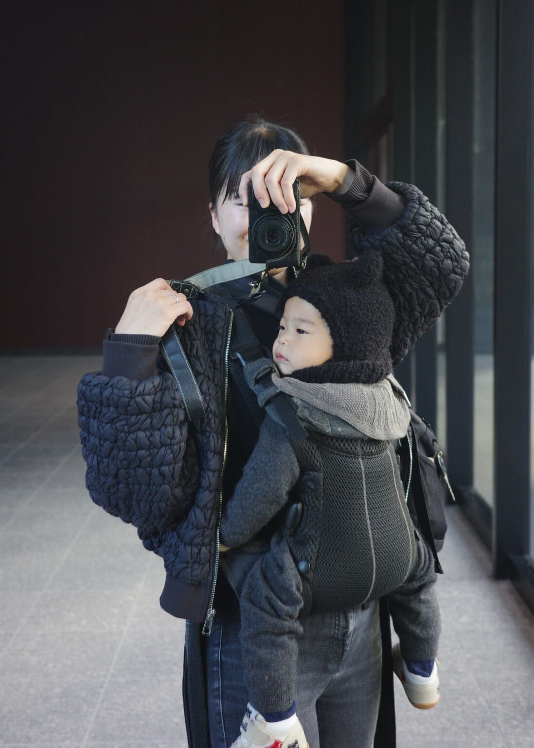 Person taking a photo with a camera, carrying a child in a front-facing carrier. Both are dressed in dark, cozy clothing.