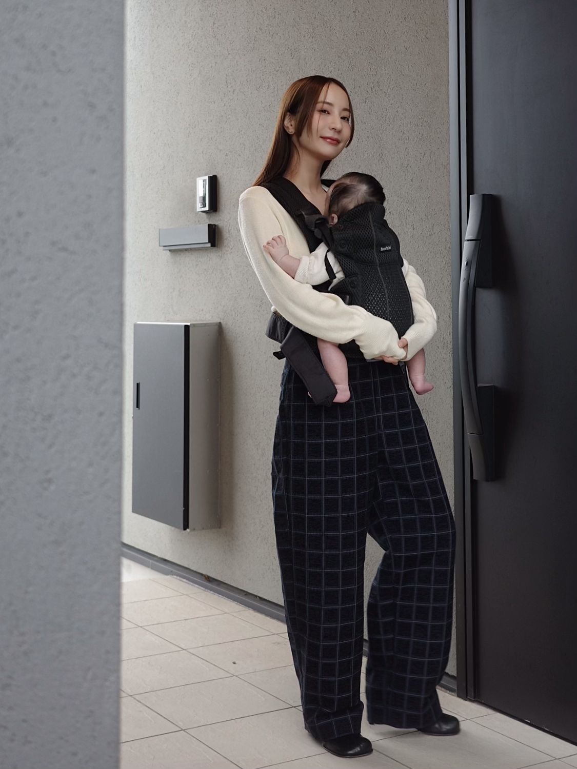 A woman stands outside, smiling, wearing a cream sweater and plaid pants, carrying a baby in a black carrier.