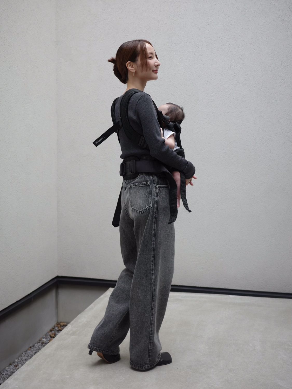 A woman stands sideways, wearing a baby carrier with a baby inside. She is dressed in a dark sweater and gray jeans against a plain background.