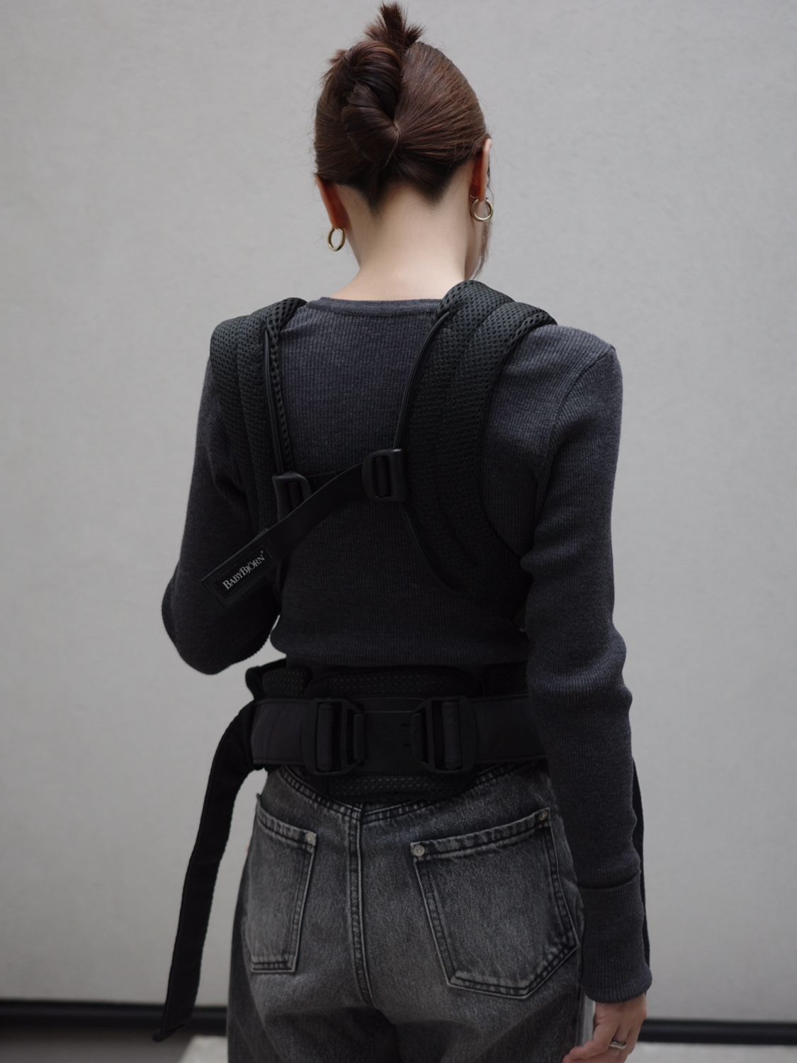 Person with a bun hairstyle wearing a dark sweater and jeans, seen from the back, with a black harness strapped over their shoulders.