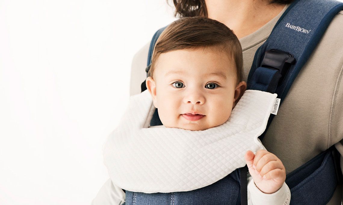 A baby with dark hair sits in a blue carrier, wearing a white bib, held by an adult. The background is white.