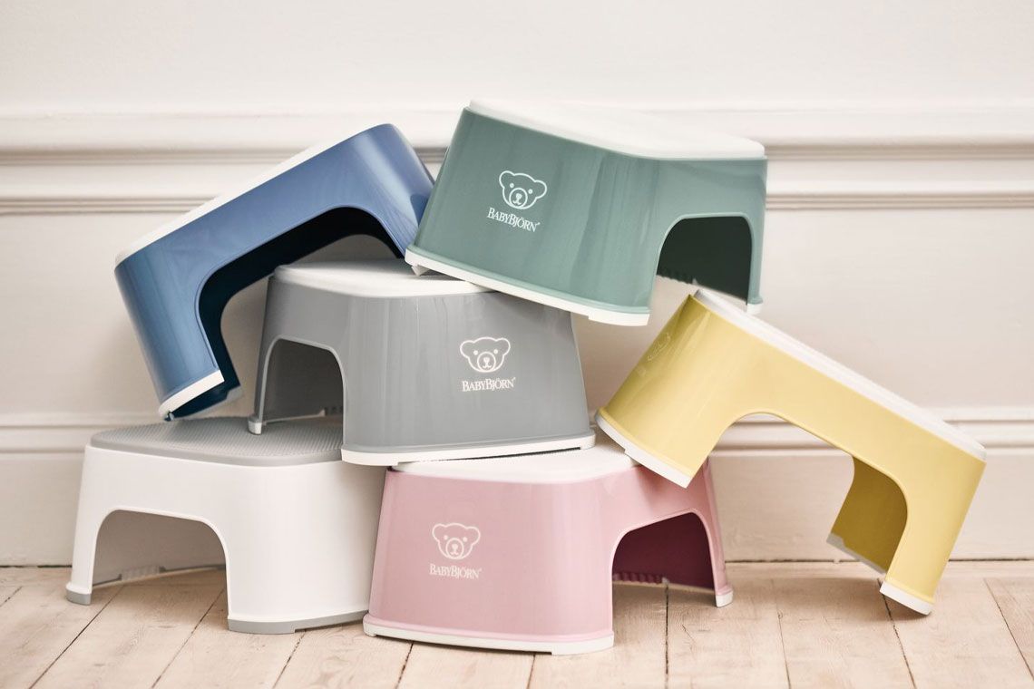 A stack of colorful plastic step stools in blue, green, yellow, pink, gray, and white, arranged against a white wall on a wooden floor.