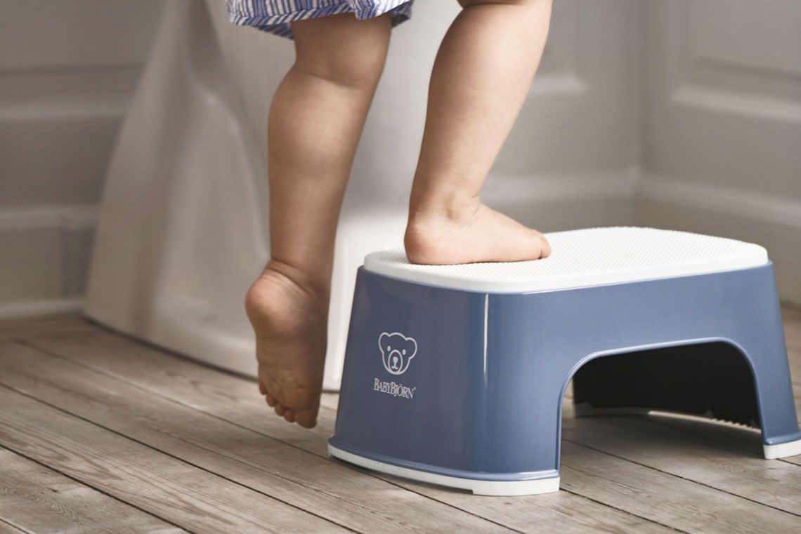 Child standing on a blue step stool in a bathroom, reaching up on tiptoes on a wooden floor.