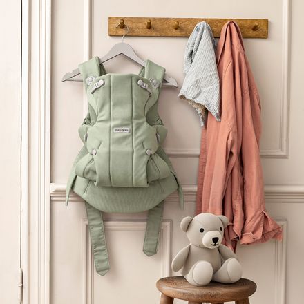 A green baby carrier hangs on a hook beside clothes, with a gray teddy bear on a stool beneath, set against a white paneled wall.