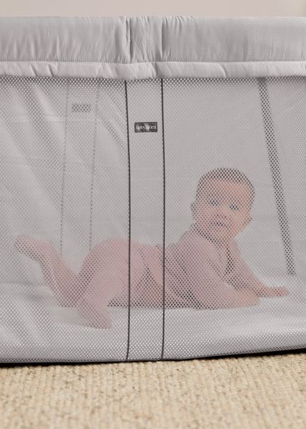 Baby lying on their stomach inside a gray mesh travel crib on a beige carpet, looking towards the camera.