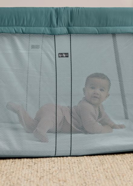 Baby lying inside a green mesh travel crib, looking out through the mesh sides, with a carpeted floor underneath.