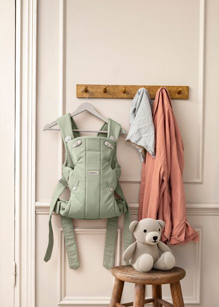 A mint green baby carrier hangs on a wall alongside pastel clothes, with a beige teddy bear on a wooden stool below.