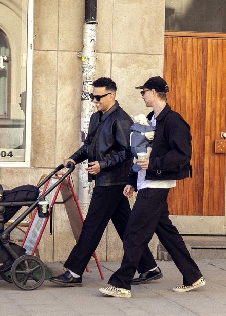 Two men walking on a sidewalk; one pushing a stroller, the other carrying a baby in a front carrier, both wearing dark clothing and sunglasses.