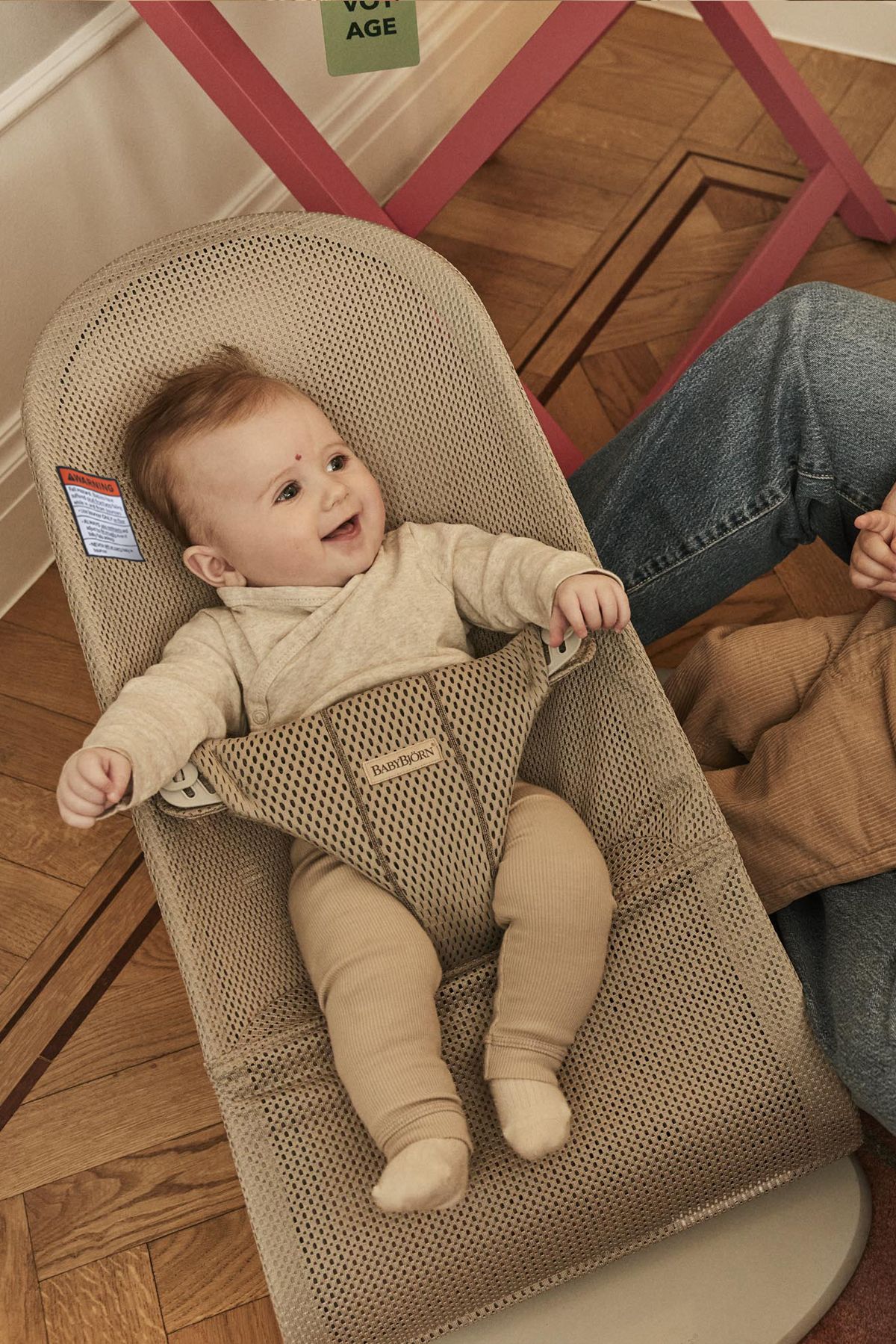 A smiling baby in a beige bouncer seat, wearing a light outfit, with an adult's arm visible nearby on a wooden floor.