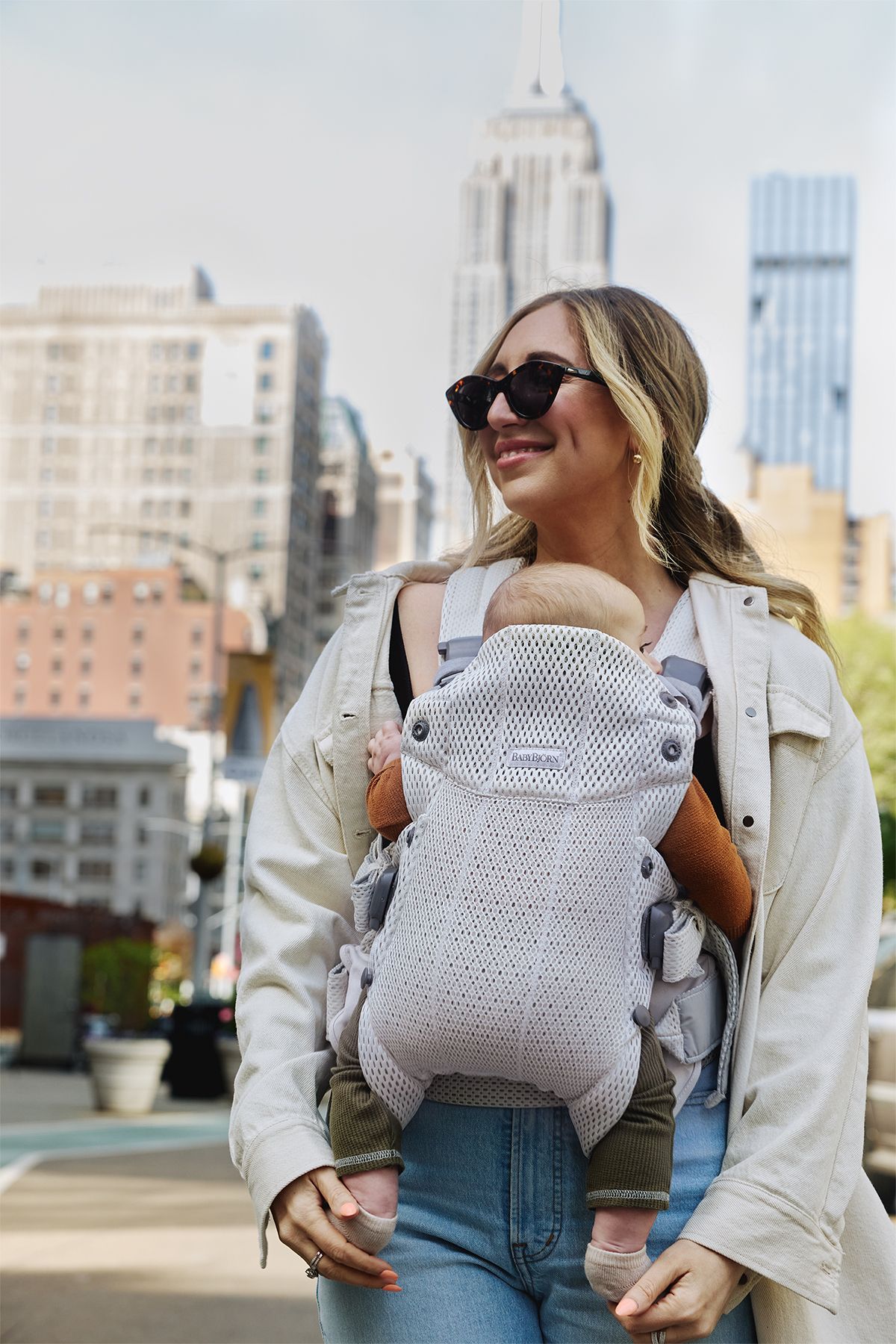 A woman wearing sunglasses carries a baby in a gray carrier. They are outdoors in a city with tall buildings in the background.
