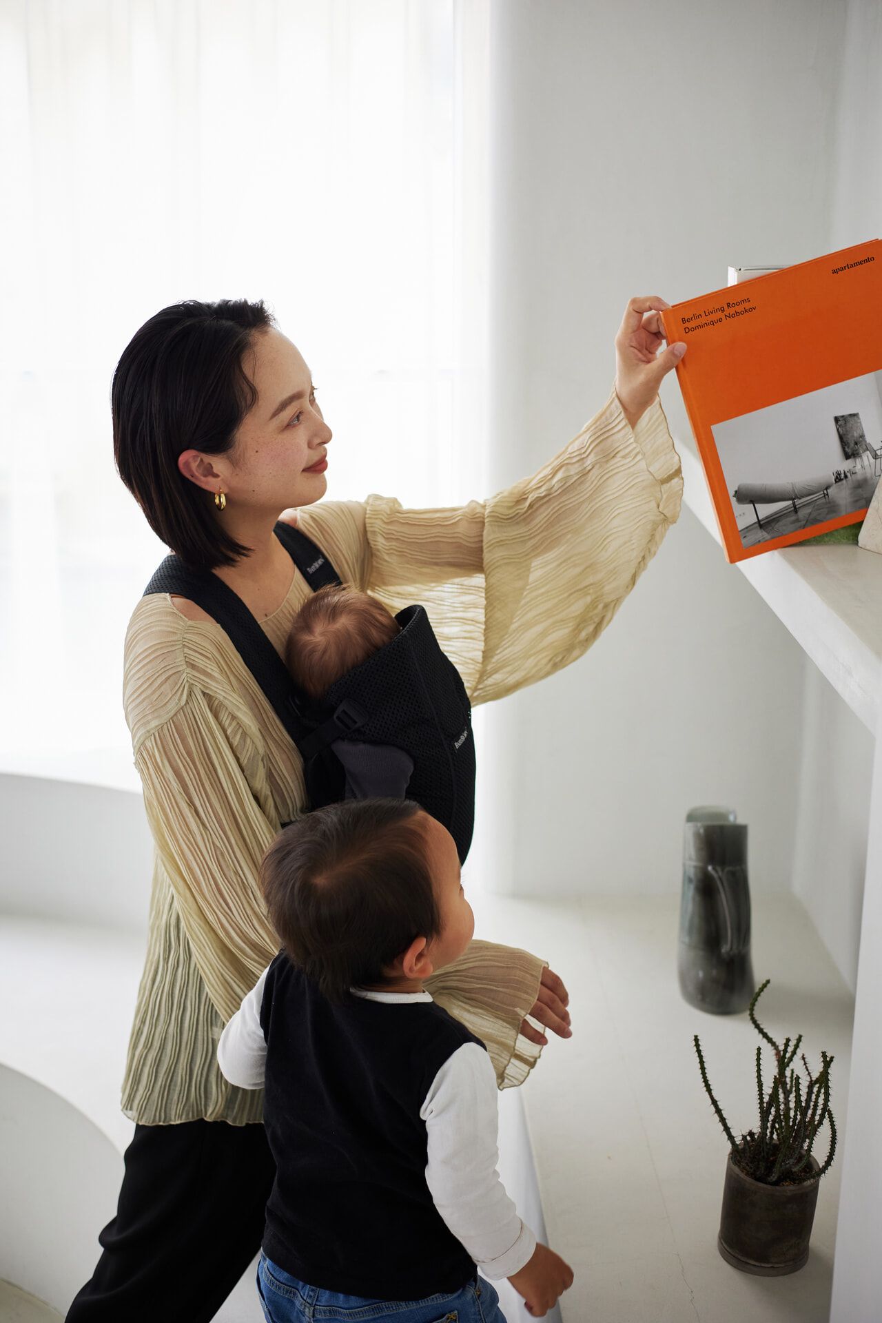 A woman holds a book while carrying a baby in a sling. A young child stands beside her, looking at the book. Bright, minimalistic room.