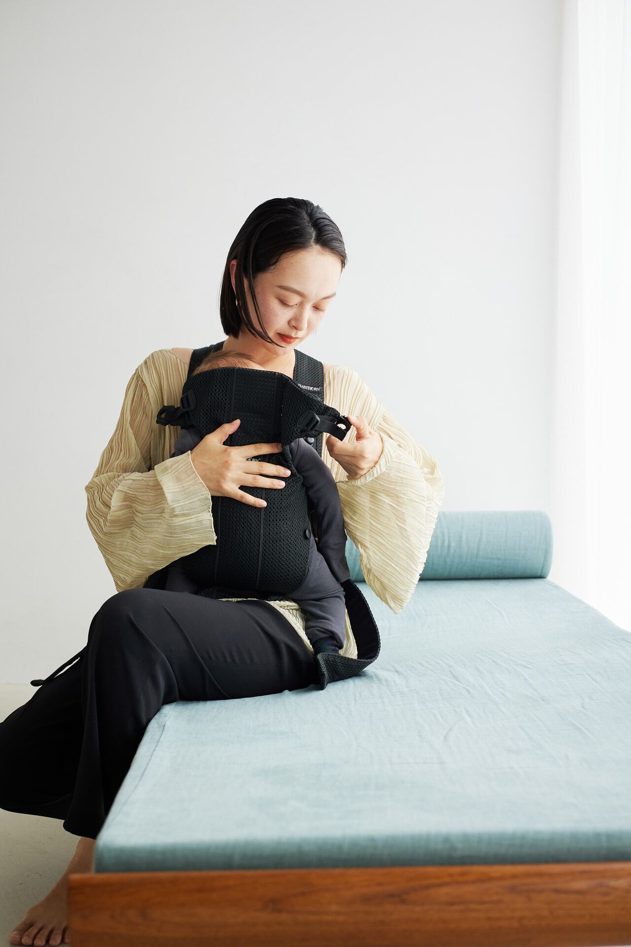 Woman sitting on a daybed, adjusting a baby carrier with a baby inside, wearing a beige top and black pants in a bright room.