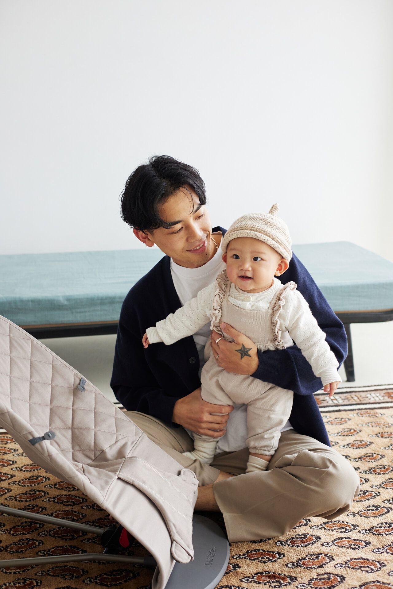 A man sits on the floor, smiling at a baby dressed in white and beige, who stands with support. A baby seat is nearby on a patterned rug.