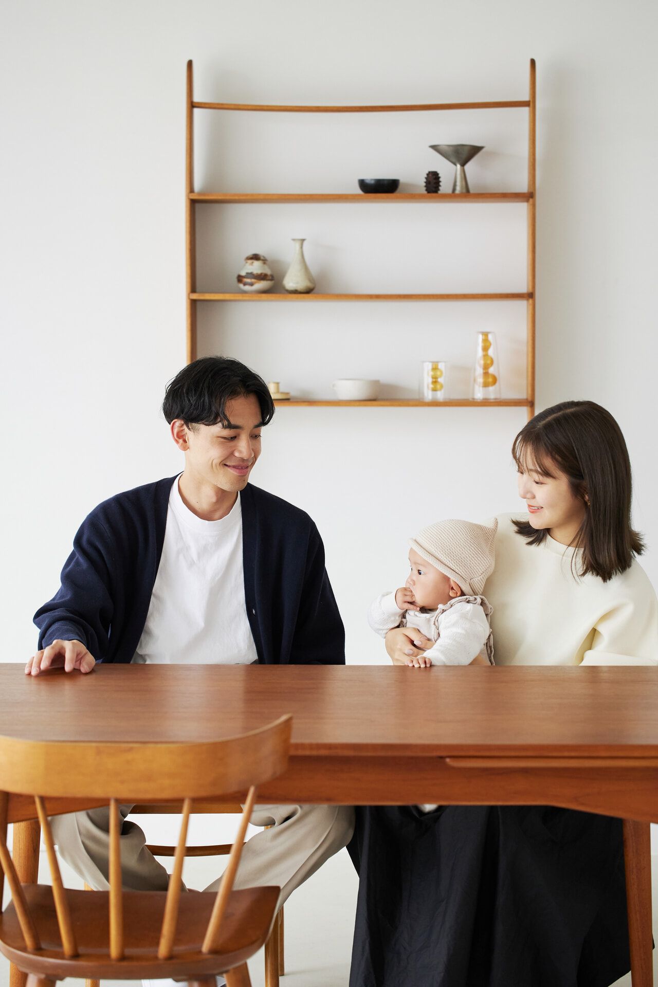 A smiling couple sits at a wooden table, with the woman holding a baby wearing a knit hat. A minimalist shelf is in the background.