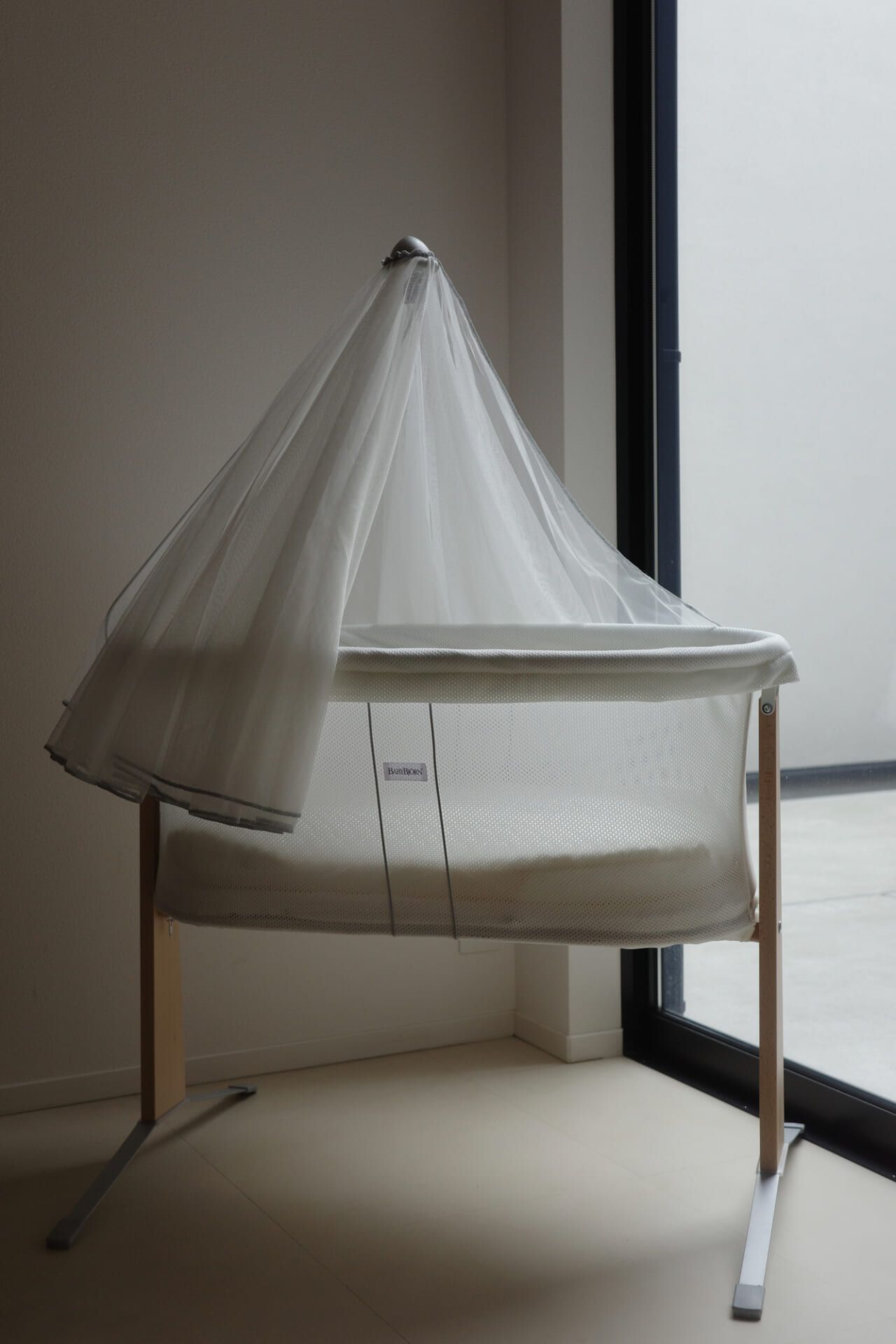 A minimalist baby bassinet with a sheer canopy stands by a large window, allowing natural light to illuminate its modern design.