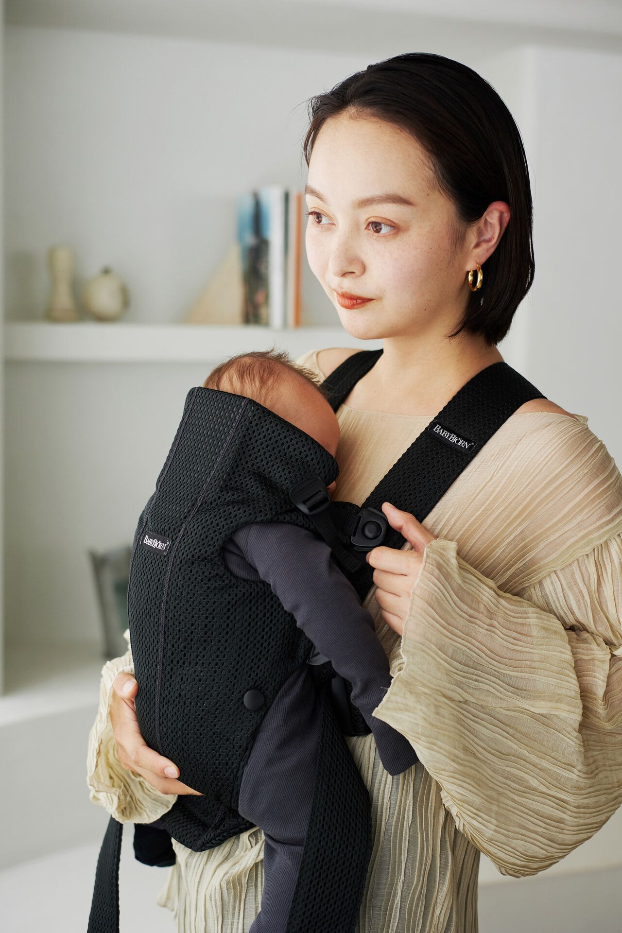 A woman holds a baby in a black carrier. She wears a beige dress and stands in a softly lit room with shelves in the background.