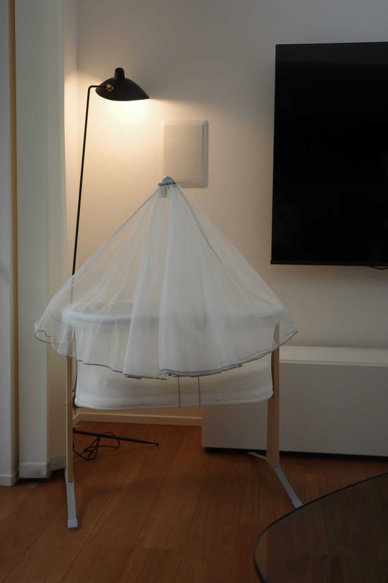 A white bassinet with a sheer canopy is placed near a floor lamp and a wall-mounted TV on a wooden floor.
