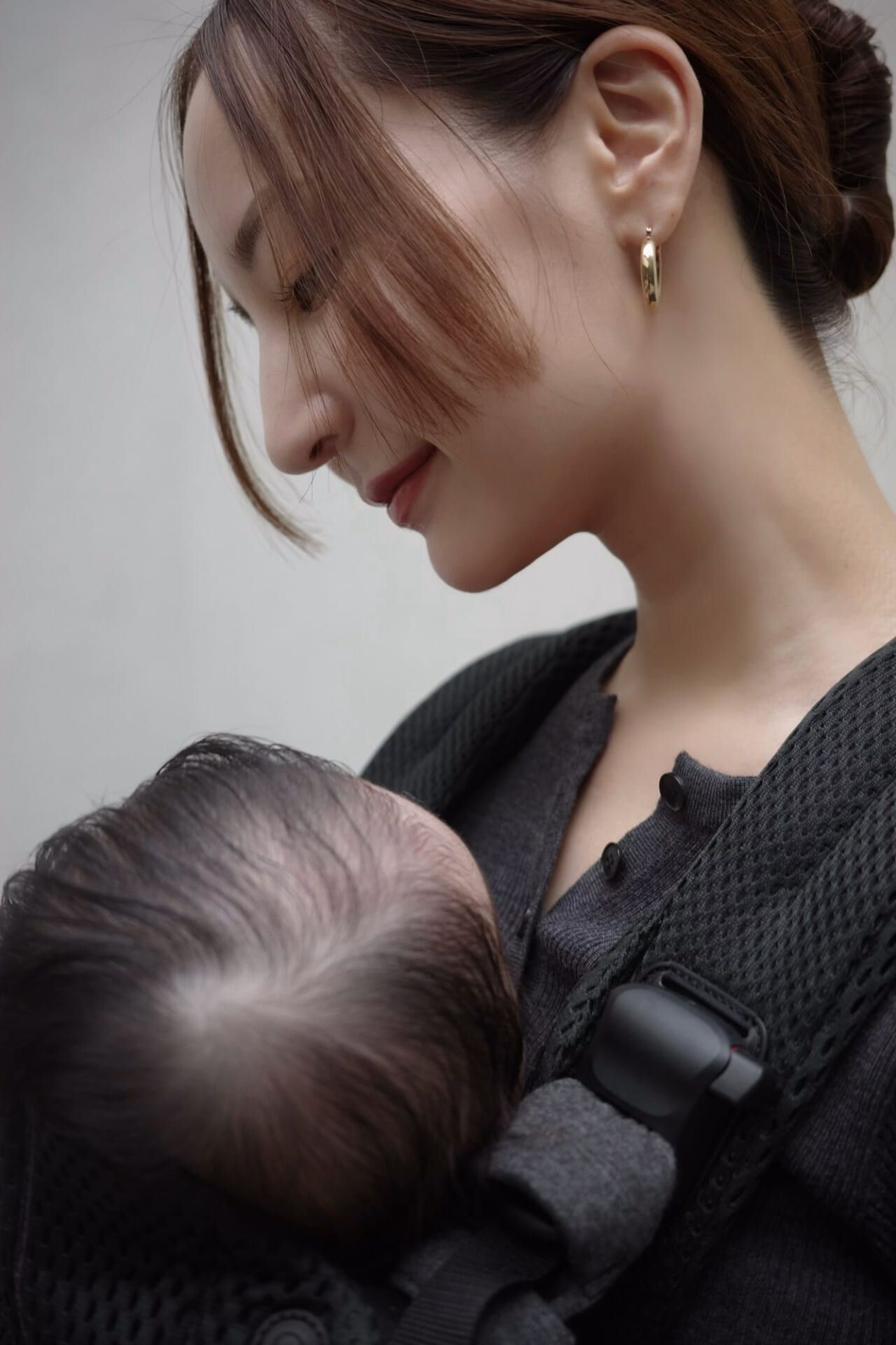 A woman gently gazes at a baby in a carrier, wearing a dark outfit and gold earring, with her hair tied back.